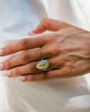 A close-up of a hand wearing a pear-shaped yellow lab diamond ring with a halo of small clear stones.